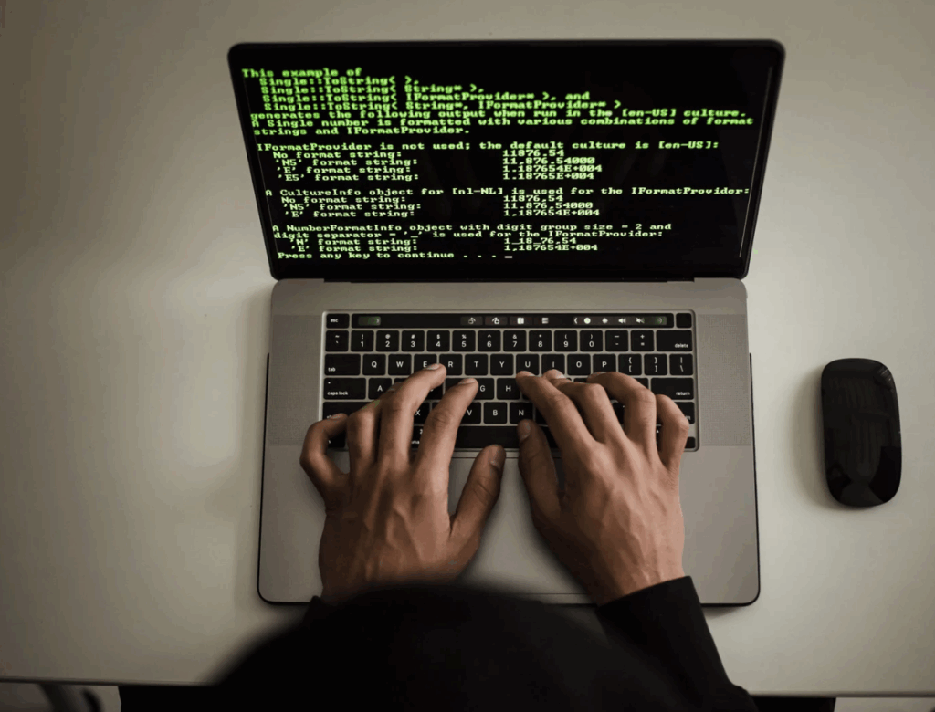 A person in a dark hoodie typing on a laptop with green text code displayed on the screen, suggesting a cybersecurity breach or hacking activity.