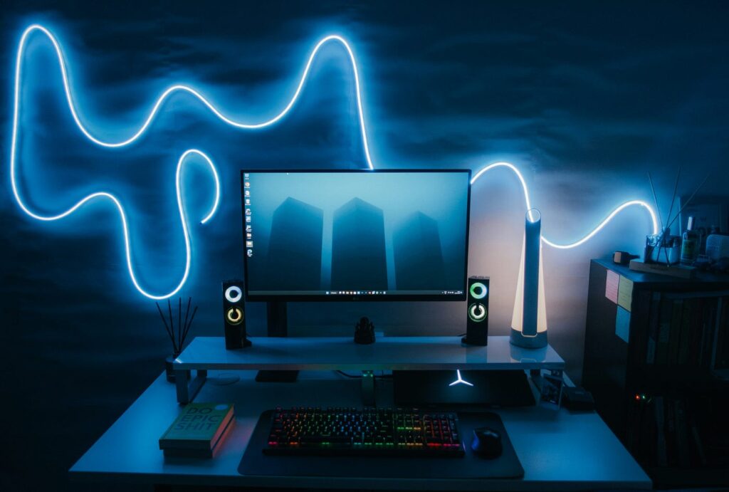 A modern computer desk setup with a glowing blue neon light on the wall behind a widescreen monitor, speakers with LED rings, a white tower fan, a rainbow backlit keyboard, and a shelf of books to the side.